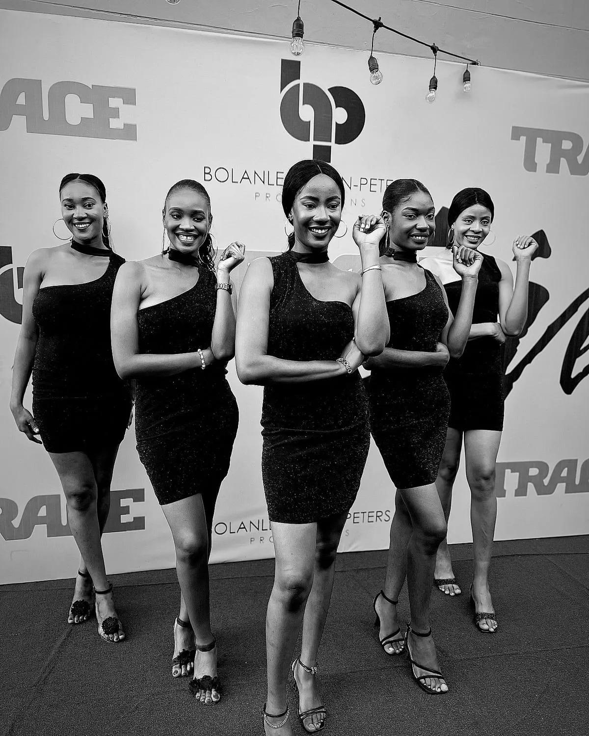 Five Anara ushers in matching black dresses posing in front of an event press wall