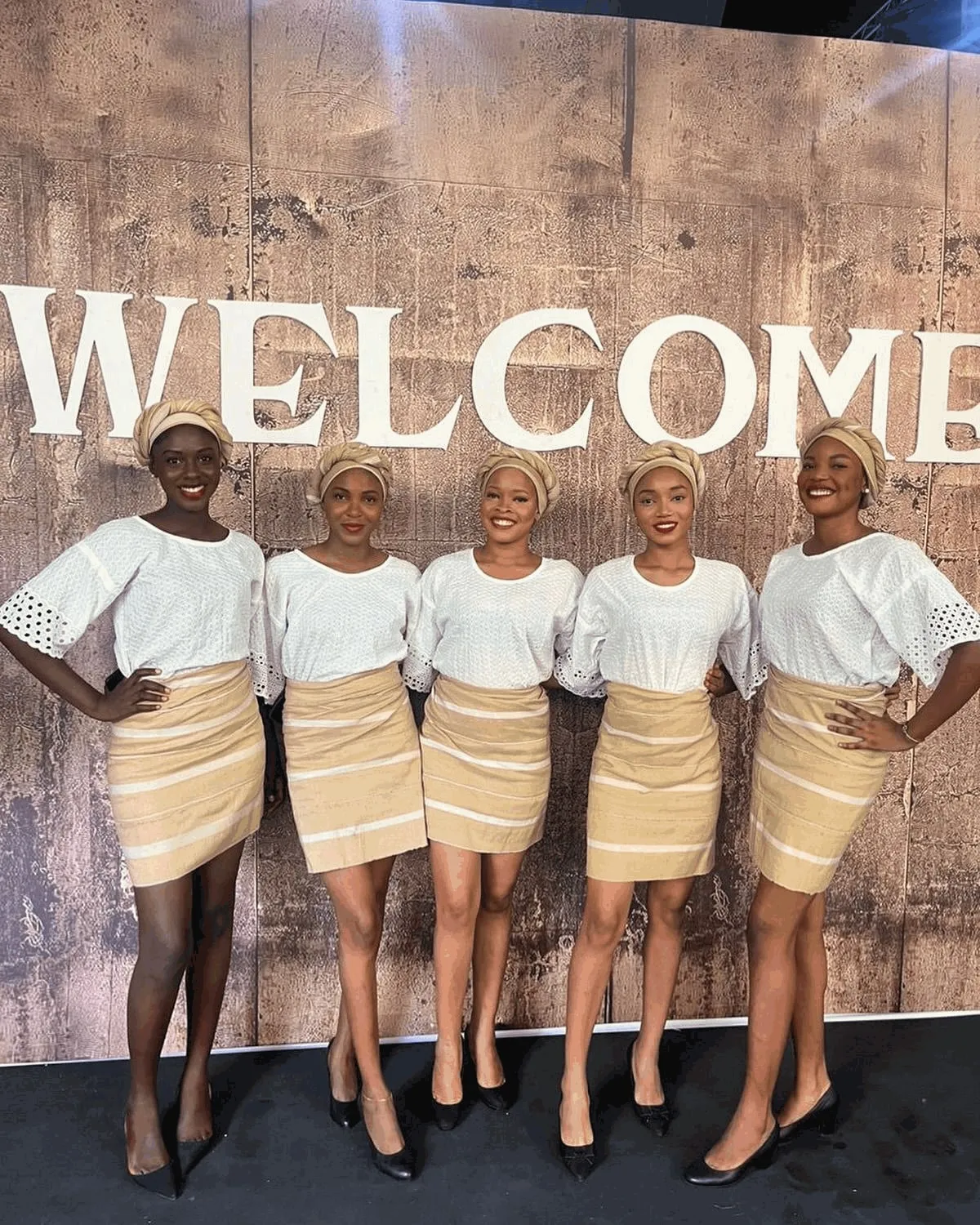 Five Anara ushers in white tops, striped skirts and matching head wraps in front of a welcome wall